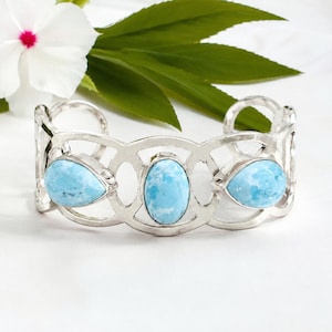 May include: A silver cuff bracelet featuring an openwork metal design with three light blue, marbled stones. The centre stone is oval-shaped, while the two flanking stones are pear-shaped. The metal band has a polished, reflective finish.