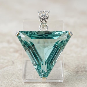 May include: A triangular-cut, translucent teal gemstone pendant featuring a faceted surface. The stone is set in a polished silver-toned metal bail with an intricate, openwork filigree pattern at the top, designed for attachment to a necklace chain.