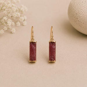 May include: A pair of gold drop earrings featuring rectangular, textured, deep red stone pendants. Each earring is suspended from a simple gold hook wire, showcasing a vertical design with a rough, crystalline surface texture on the red stones.