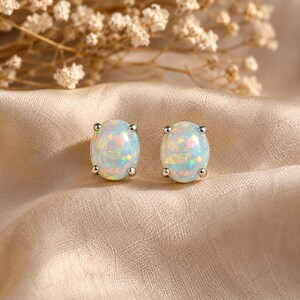 May include: A pair of oval-shaped stud earrings featuring iridescent white opals set in silver-toned metal prongs. Each earring displays a smooth, rounded surface with flashes of blue, green, and pink light reflecting from within the stones.