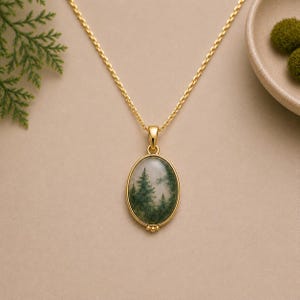 Pode incluir: Um colar de ouro com um pendente oval de ágata musgosa. A pedra apresenta inclusões verdes naturais que lembram uma paisagem florestal. O pendente está suspenso numa delicada corrente de ouro e tem três pequenas contas douradas na base.