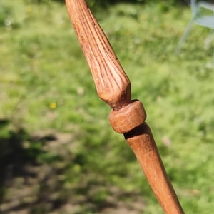 Peut inclure: Une baguette en bois sculptée à la main, avec une pointe et un motif texturé et strié. La baguette est de couleur marron chaud et comporte une section arrondie près du centre. L'arrière-plan est flou, avec de l'herbe verte et un soupçon de chaise.