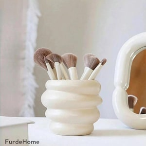 Puede incluir: Un soporte para brochas de maquillaje de cerámica color crema con un diseño apilado único. El soporte contiene varios pinceles de maquillaje con mangos blancos y cerdas marrones suaves. Un espejo blanco está en el fondo.