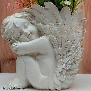 Könnte beinhalten: Ein weißer Engel-Blumentopf mit detaillierten Federflügeln, der seine Knie umarmt und die Augen geschlossen hat. Der Topf ist mit rosa, weißen und grünen Kunstblumen gefüllt. Der Text "FurdeHome" ist sichtbar.