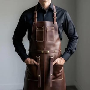 May include: A brown leather apron with two chest pockets and two large front pockets. The apron has adjustable straps and brass-colored rivets. The person in the image is wearing a black long-sleeve shirt.