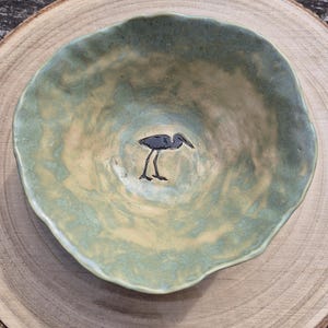 May include: A round, shallow ceramic bowl with an uneven, wavy rim. The interior features a mottled, sage green and cream-coloured glaze. A dark, detailed silhouette of a long-legged wading bird is centred at the bottom of the bowl.