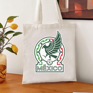 Peut inclure: Un sac fourre-tout blanc avec l'embl&egrave;me national mexicain. L'embl&egrave;me est un motif vert, blanc et rouge avec un aigle central tenant un serpent et le mot "M&Eacute;XICO" en dessous. Le sac a une forme simple et rectangulaire.