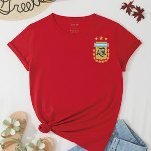 May include: A red short-sleeved t-shirt featuring a crew neckline and a graphic on the left chest. The graphic displays the AFA logo in gold, light blue, and white, topped with three gold stars and surrounded by a gold laurel wreath design.