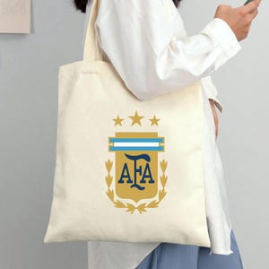 May include: A cream-colored canvas tote bag featuring a centered graphic of the Argentina Football Association crest. The design includes three gold stars above a shield with the letters AFA in dark blue, flanked by gold laurel branches.