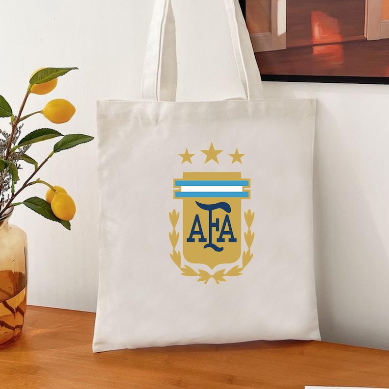 May include: A white canvas tote bag featuring the AFA logo in gold, light blue, and dark blue, topped with three gold stars. The design includes a central shield with the letters AFA, flanked by gold laurel branches on a plain white fabric background.