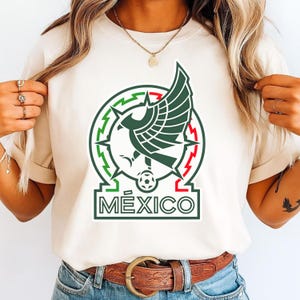Peut inclure: T-shirt cr&egrave;me avec l'embl&egrave;me national mexicain. L'embl&egrave;me comprend un motif circulaire vert et rouge avec un aigle stylis&eacute;, un ballon de football et le mot "M&Eacute;XICO" en vert. Le t-shirt est port&eacute; par une personne.