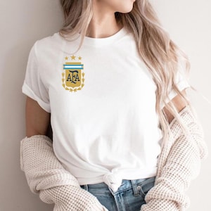 May include: A white short-sleeved t-shirt featuring a graphic on the left chest. The graphic displays the letters AFA in dark blue, topped with a light blue and white horizontal bar, three gold stars, and a gold laurel wreath design.