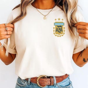 May include: A cream-colored short-sleeve t-shirt featuring a small graphic on the left chest. The graphic displays a gold shield with the letters AFA in dark blue, topped with three gold stars and surrounded by gold laurel leaf accents.