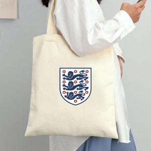 Puede incluir: Una bolsa de tela color crema con un emblema de escudo centrado. El escudo muestra tres leones azul oscuro apilados con lenguas y garras rojas, rodeados de peque&ntilde;os detalles florales rojos, todo ello encerrado dentro de un borde azul oscuro.