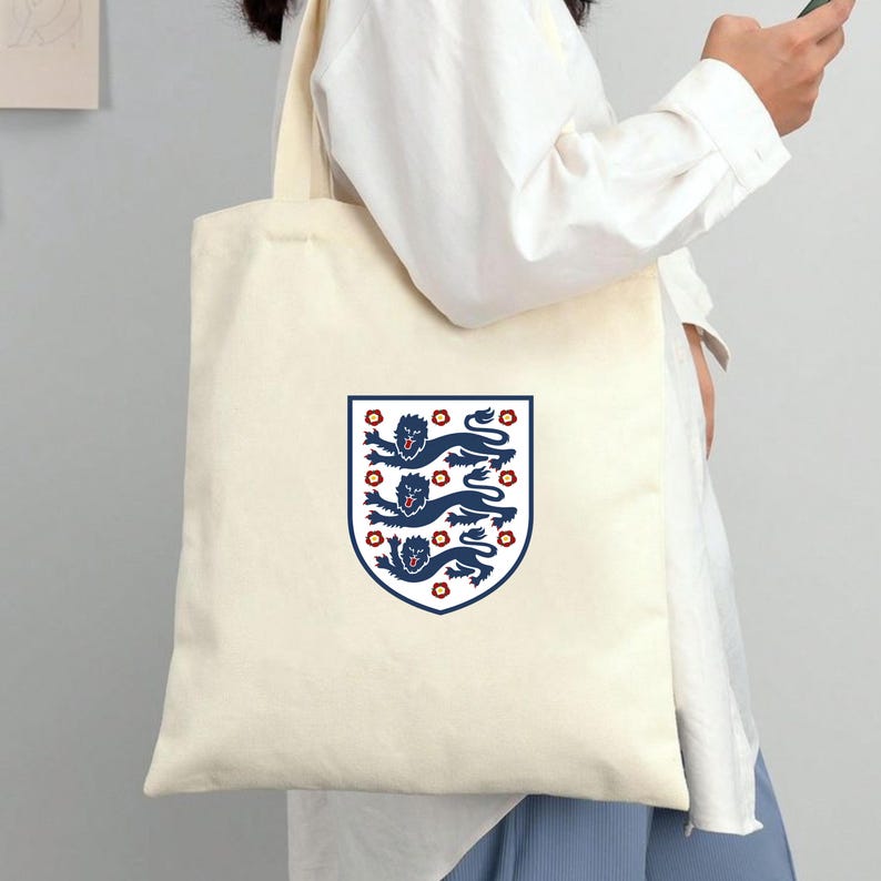 Puede incluir: Una bolsa de tela color crema con un emblema de escudo centrado. El escudo muestra tres leones azul oscuro apilados con lenguas y garras rojas, rodeados de peque&ntilde;os detalles florales rojos, todo ello encerrado dentro de un borde azul oscuro.
