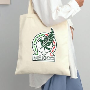Peut inclure: Sac fourre-tout cr&egrave;me avec l'embl&egrave;me national mexicain en vert, rouge et blanc. L'embl&egrave;me comprend un aigle avec un serpent, un ballon de football et le mot "M&eacute;xico". Le sac est port&eacute; par une personne v&ecirc;tue d'une chemise blanche et d'un pantalon bleu.