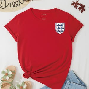 Puede incluir: Una camiseta roja de manga corta con cuello redondo y un peque&ntilde;o emblema en forma de escudo en el pecho izquierdo. El emblema muestra tres leones azules apilados verticalmente sobre un fondo blanco. La etiqueta interior dice SHEIN.