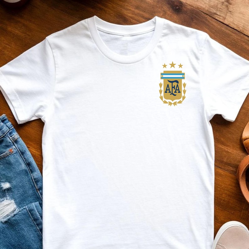 May include: A white short-sleeved t-shirt featuring a small graphic on the left chest. The graphic displays the AFA logo in gold and blue, topped with three gold stars and framed by gold laurel branches on a white background.