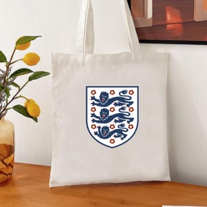 Puede incluir: Una bolsa de tela color crema que presenta un emblema de escudo centrado. El escudo muestra tres leones azul oscuro apilados con lenguas y garras rojas, rodeados de peque&ntilde;os detalles florales rojos, todo encerrado en un borde azul oscuro.