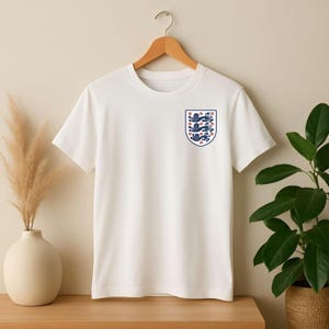 Puede incluir: Una camiseta blanca de manga corta con un peque&ntilde;o escudo en el pecho izquierdo. El escudo tiene forma de blas&oacute;n y contiene tres leones azules apilados con lenguas y garras rojas, delineados en azul, representando al equipo nacional de Inglaterra.