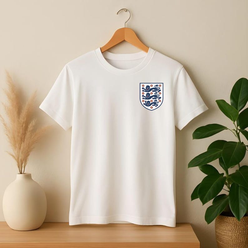 Puede incluir: Una camiseta blanca de manga corta con un peque&ntilde;o escudo en el pecho izquierdo. El escudo tiene forma de blas&oacute;n y contiene tres leones azules apilados con lenguas y garras rojas, delineados en azul, representando al equipo nacional de Inglaterra.