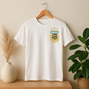 May include: A white short-sleeved t-shirt featuring a gold and blue crest on the left chest. The crest displays the letters AFA in dark blue, topped with three gold stars and a light blue horizontal bar, all framed by gold laurel leaves.