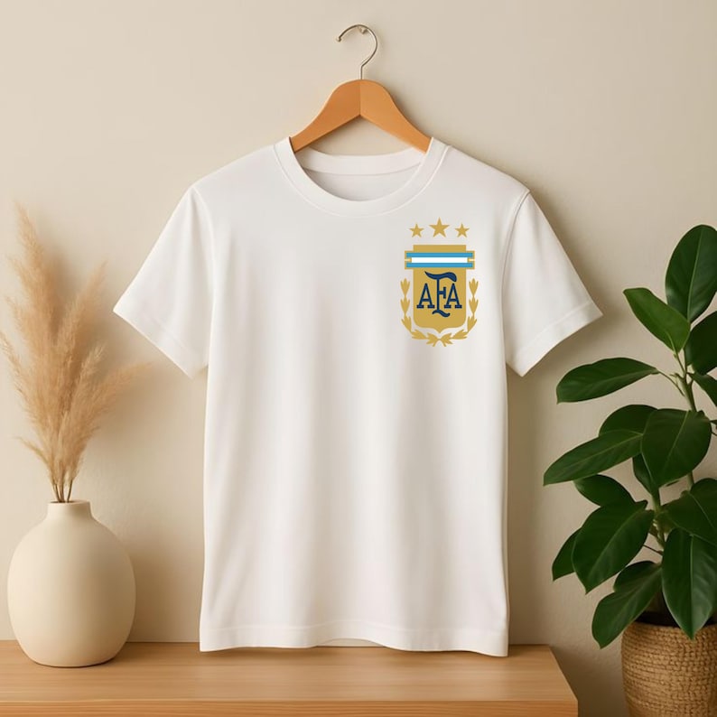 May include: A white short-sleeved t-shirt featuring a gold and blue crest on the left chest. The crest displays the letters AFA in dark blue, topped with three gold stars and a light blue horizontal bar, all framed by gold laurel leaves.