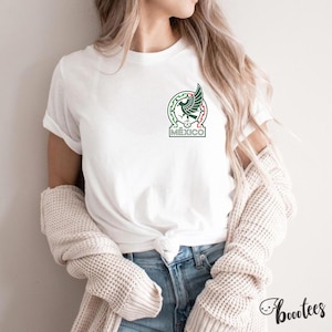 Peut inclure: T-shirt blanc avec les armoiries mexicaines, dans un design vert, rouge et blanc. Le t-shirt a un col rond et des manches courtes. Le mot "M&Eacute;XICO" est imprim&eacute; sous l'embl&egrave;me. Le t-shirt est port&eacute; avec un jean bleu.