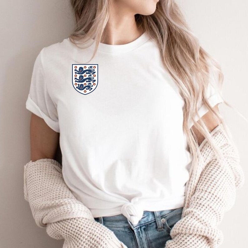 Puede incluir: Una camiseta blanca de manga corta con un peque&ntilde;o escudo en el pecho izquierdo. El escudo tiene forma de blas&oacute;n y contiene tres leones azules apilados con lenguas y garras rojas, rodeados por diez peque&ntilde;as flores rojas sobre fondo blanco.