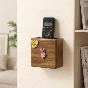 Puede incluir: Un organizador de pared de madera con un mando a distancia en su interior. El organizador está decorado con una abeja amarilla y negra y una flor rosa. El organizador es de madera marrón oscuro.