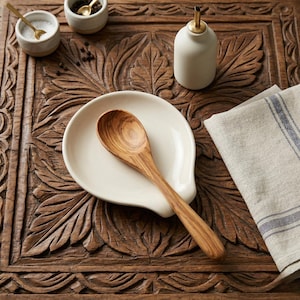 May include: A wooden spoon rests on a white ceramic spoon rest. A small white ceramic container with a gold spoon is in the background. A white ceramic oil dispenser with a gold spout is also visible. The items are on a carved wooden surface.