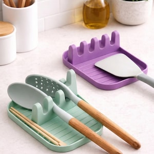 Puede incluir: Un reposa-utensilios verde claro con una cuchara ranurada, una cuchara sólida y palillos chinos. Un reposa-utensilios morado sostiene una espátula. Los utensilios tienen mangos de madera. Utensilios de cocina.