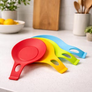 May include: Four brightly coloured silicone spoon rests in red, yellow, green, and blue. Each rest has a circular area for the spoon and a handle with a hole for hanging. The spoon rests are arranged in a slightly overlapping fan shape.