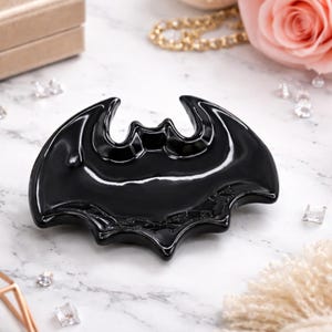 Puede incluir: Una bandeja decorativa negra brillante con forma de símbolo de murciélago. La bandeja tiene una superficie lisa y reflectante y está colocada sobre un fondo de mármol. La bandeja se utiliza probablemente para guardar joyas u objetos pequeños.
