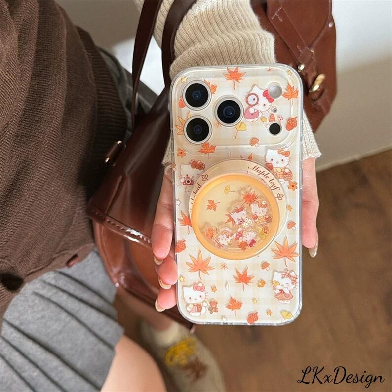 K&ouml;nnte beinhalten: Transparente Handyh&uuml;lle mit Herbstthema, mit orangefarbenen Ahornbl&auml;ttern und Hello Kitty-Figuren. Die H&uuml;lle hat einen Pop-up-Griff mit schwebendem Glitzer und dem Text "Maple leaf". Die H&uuml;lle ist f&uuml;r ein Smartphone konzipiert.
