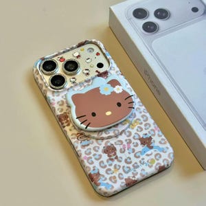 May include: A white and brown iPhone case featuring a Hello Kitty design with a pop-up grip. The case has a leopard print pattern with cartoon characters. A white iPhone box is in the background.