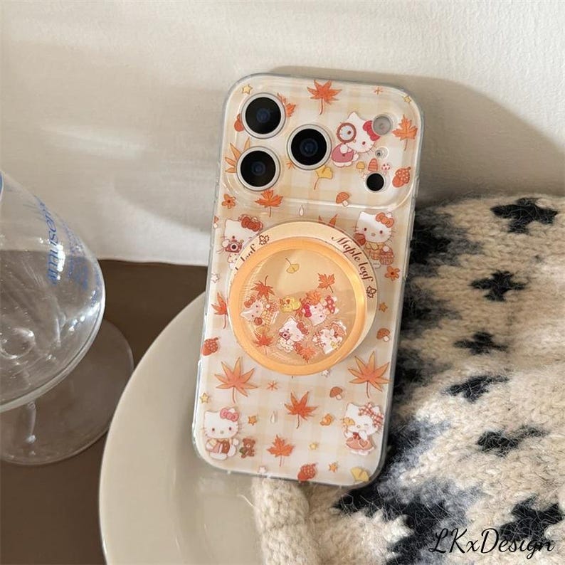 K&ouml;nnte beinhalten: Transparente Handyh&uuml;lle mit Hello Kitty-Design, Ahornbl&auml;ttern und einem Pop-up-Griff. Die H&uuml;lle hat ein hellbeige kariertes Muster und einen transparenten Griff mit schwebendem Konfetti. F&uuml;r ein Smartphone.