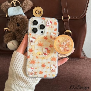K&ouml;nnte beinhalten: Transparente Handyh&uuml;lle mit Hello Kitty-Design, Herbstbl&auml;ttern und einem Vichy-Muster. Die H&uuml;lle wird in der N&auml;he einer braunen Ledertasche und eines Teddyb&auml;r-Schl&uuml;sselanh&auml;ngers gehalten. Ein rundes, transparentes Zubeh&ouml;r mit dem Text "Maple leaf" ist ebenfalls sichtbar.