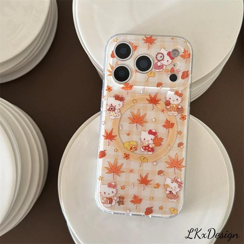 K&ouml;nnte beinhalten: Transparente Handyh&uuml;lle mit einem sich wiederholenden Muster aus Hello Kitty-Charakteren, Ahornbl&auml;ttern und Sternen in Orange und Wei&szlig;. Die H&uuml;lle hat einen transparenten Rand und einen kreisf&ouml;rmigen Ausschnitt. Das Design ist vor einem hellorange-wei&szlig; karierten Hintergrund angeordnet.