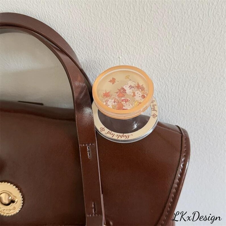 K&ouml;nnte beinhalten: Eine braune Lederhandtasche mit goldenem Verschluss und Griff. Auf der Tasche befindet sich ein kleiner, runder Beh&auml;lter mit durchsichtigem Deckel und herbstlichen Dekorationen. Der Beh&auml;lter hat einen goldenen Rand und die Worte "Maple leaf" sind auf dem Rand aufgedruckt.