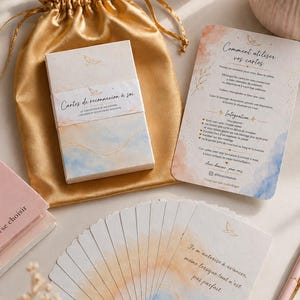 Peut inclure: Un jeu de cartes de reconnexion à soi avec des motifs aquarelle, dans une pochette dorée. La boîte et les cartes individuelles présentent des teintes douces de bleu et d'orange, avec des accents dorés et du texte. Une bougie et un stylo sont également visibles.