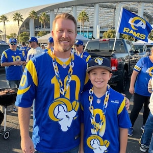 Könnte beinhalten: Zwei Personen tragen blau-gelbe Trikots der Los Angeles Rams mit der Nummer 10 und einem Widderkopf-Logo. Beide tragen weiß-blaue Kunststoffketten mit einem großen Widderkopf-Anhänger. Eine Person trägt eine dunkelblaue Baseballkappe mit Widder-Logo.