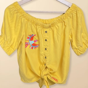 Może przedstawiać: Jasnożółty top z odkrytymi ramionami, krótkimi rękawami i wiązanym przodem. Koszula posiada kolorowy haftowany wzór ptaka z przodu i jest zapinana na guziki pośrodku. Nazwa marki "ZABIEL" jest widoczna w prawym dolnym rogu.