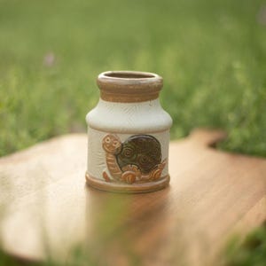 Może przedstawiać: Biały ceramiczny kubek z czarnym uchwytem i napisem "But First, Coffee" czarną, pogrubioną czcionką. Kubek stoi na białej powierzchni. Kubek ma około 10 cm wysokości.