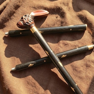 Può includere: Bastone decorativo con manico a forma di testa di coniglio intagliata e fusto in legno scuro. Il manico è marrone e il fusto è nero con dettagli dorati. Il bastone è appoggiato su un tessuto marrone.
