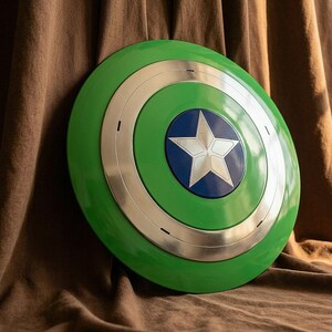 Pode incluir: Um escudo verde e prateado com um círculo azul central e uma estrela branca. O escudo tem um design circular com anéis concêntricos verdes, prateados e azuis. É um adereço ou item de colecionador.
