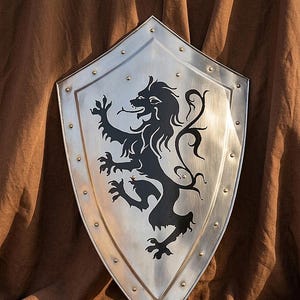 Może przedstawiać: Srebrna tarcza z czarnym herbem lwa. Tarcza ma spiczaste dno i jest obramowana nitami. Lew jest przedstawiony w stylu heraldycznym. Tło to brązowa tkanina, tworząca kontrast z metalową powierzchnią tarczy.