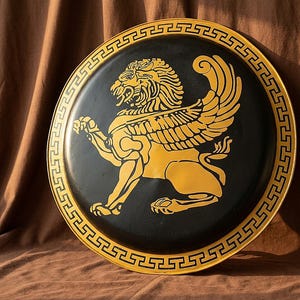 Puede incluir: Una bandeja decorativa redonda y negra con un diseño de león alado dorado. El león está en posición agachada con la boca abierta. La bandeja tiene un borde dorado con un patrón geométrico repetitivo.
