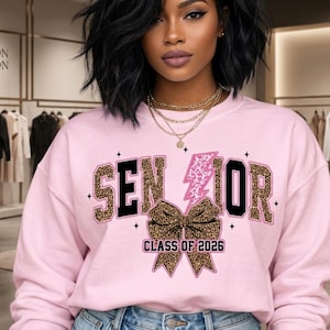 Op de afbeelding: Een lichtroze sweatshirt met het woord "SENIOR" in een mix van luipaardprint en zwarte blokletters. Een roze bliksemschicht en een luipaardprint strik zijn ook afgebeeld. De tekst "CLASS OF 2026" staat onder de strik.