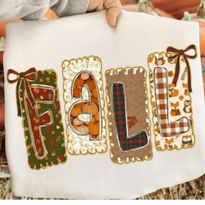 May include: A white t-shirt featuring the word FALL in large block letters. Each letter is framed by a scalloped border and decorated with seasonal patterns including pumpkins, plaid, autumn leaves, acorns, foxes, and owls, with bows on the F and L.